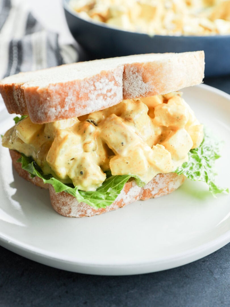 A close-up of an curried chicken salad sandwich with leafy lettuce on white bread, served on a white plate. In the background, a bowl of salad sits beside a dish of curried chicken salad for added variety.