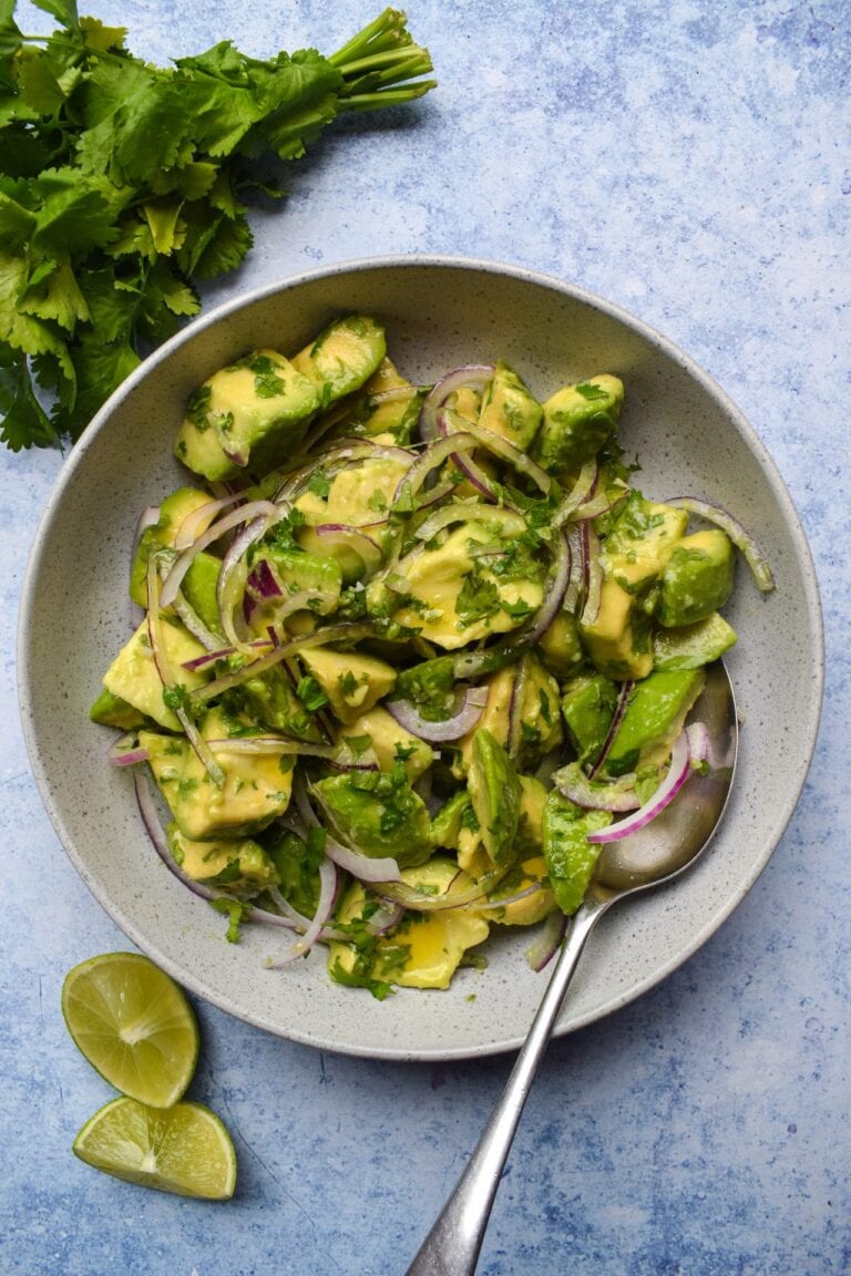 A bowl of avocado salad with red onion slices and chopped cilantro, served with lime wedges on the side. A bunch of fresh cilantro is placed above the bowl on a blue textured surface.