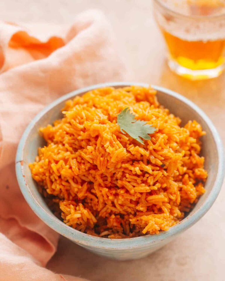 A bowl of vibrant orange-colored rice garnished with a single cilantro leaf, placed on a light surface beside a peach-colored cloth and a glass of yellow drink in the background.