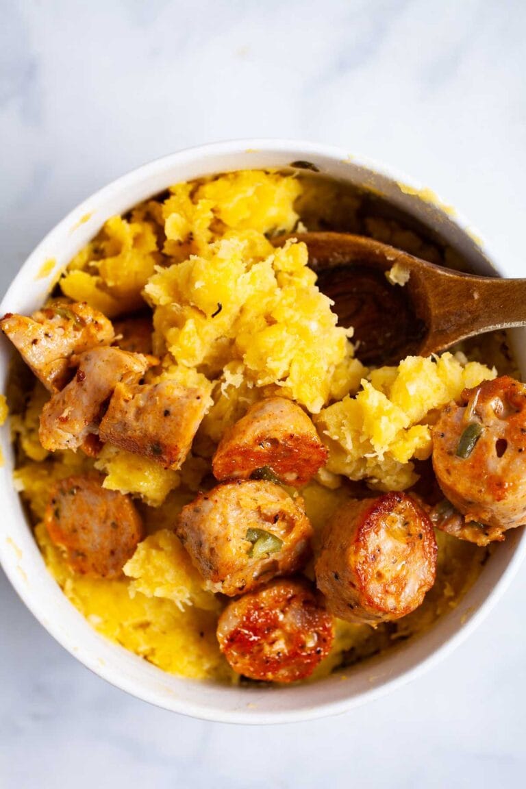 A white bowl filled with mashed yellow squash topped with sliced cooked sausage pieces, with a wooden spoon resting inside the bowl.