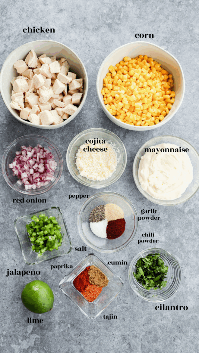 Top-down view of bowls containing mexican chicken salad ingredients: cubed chicken, corn, red onion, cotija cheese, mayonnaise, jalapeño, lime, cilantro, and spices like pepper, salt, garlic powder, chili powder, cumin, paprika, and tajin.