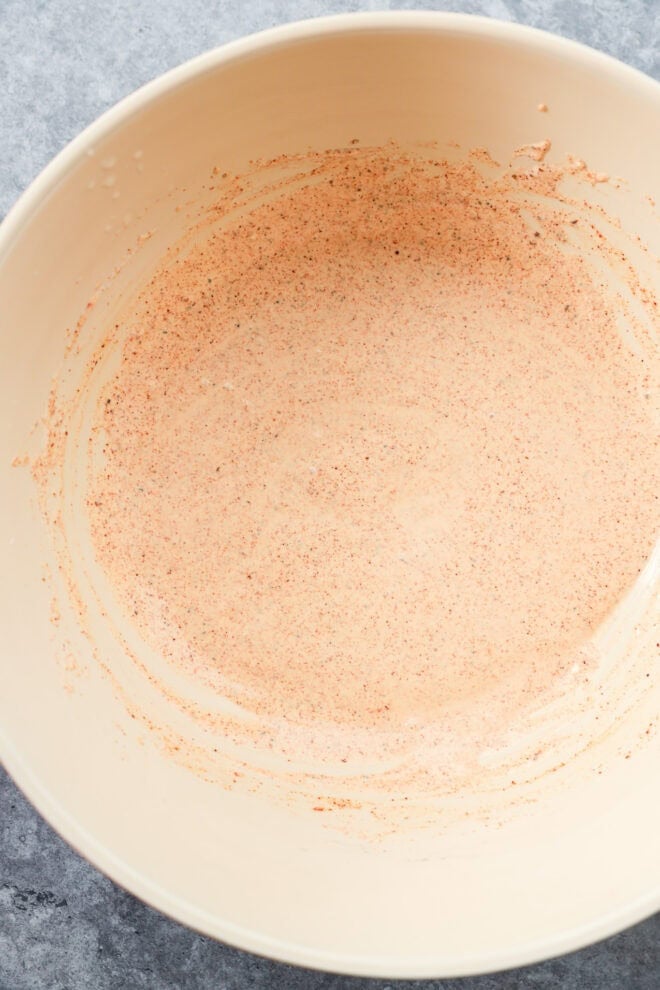 A beige mixing bowl contains a thin layer of dressing on a grey surface.