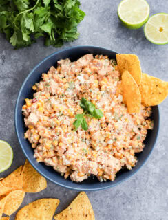 A blue bowl filled with a creamy Mexican chicken salad, garnished with cilantro and crumbled cheese, surrounded by tortilla chips. Fresh lime halves and cilantro are placed nearby on a gray surface.