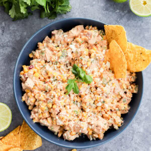 A blue bowl filled with a creamy Mexican chicken salad, garnished with cilantro and crumbled cheese, surrounded by tortilla chips. Fresh lime halves and cilantro are placed nearby on a gray surface.