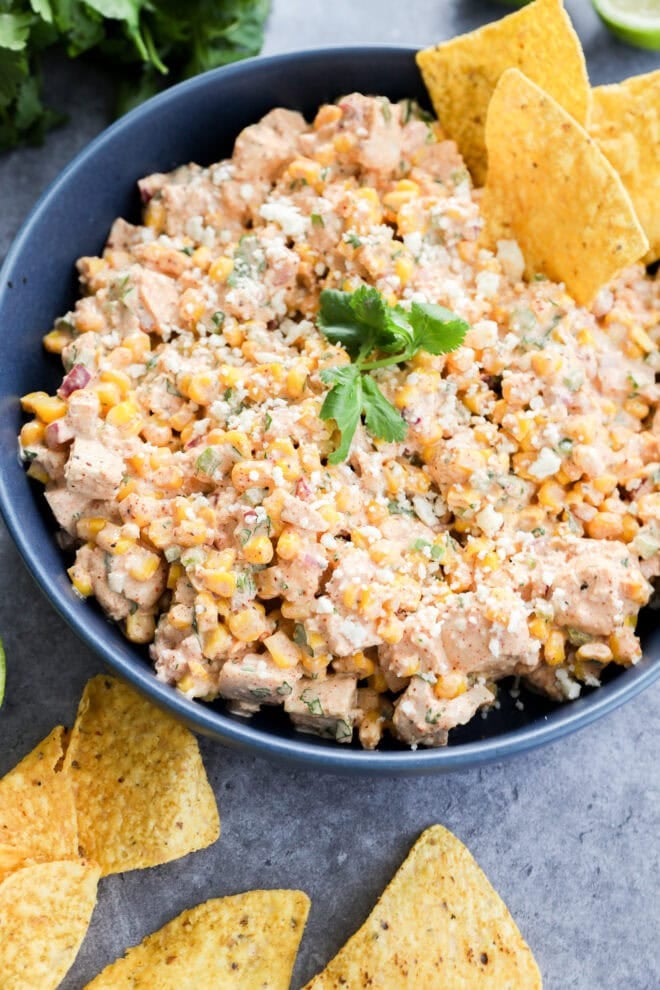 A blue bowl filled with creamy corn dip, inspired by flavors of Mexican chicken salad, topped with crumbled cheese and cilantro, surrounded by tortilla chips on a gray surface.