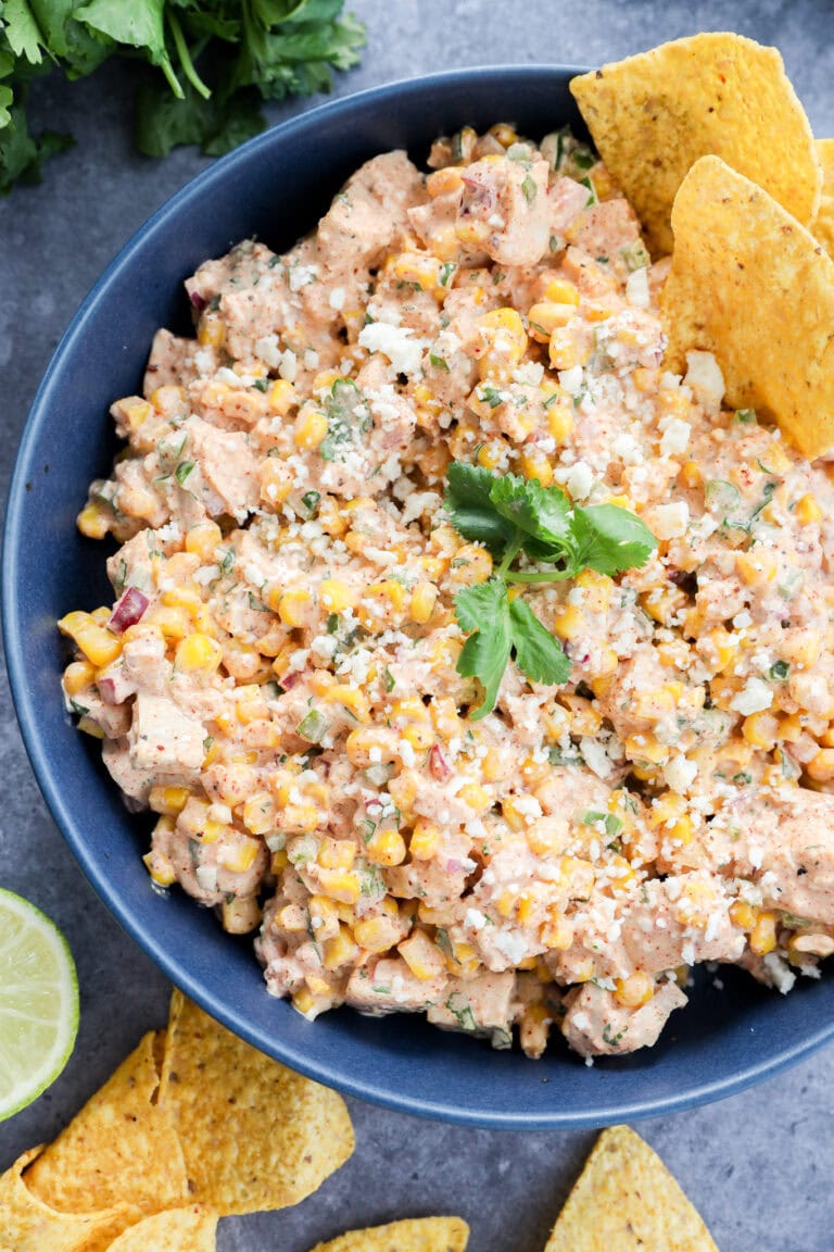 A bowl of Mexican street corn salad topped with cilantro and crumbled cheese, served with tortilla chips on the side. A lime wedge and fresh cilantro are visible nearby—perfect alongside your favorite Mexican chicken salad.