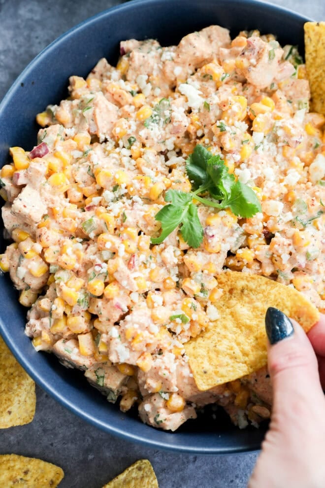 A bowl of creamy corn salad is topped with cilantro and crumbled cheese, with a hand dipping a tortilla chip into the delicious mix.