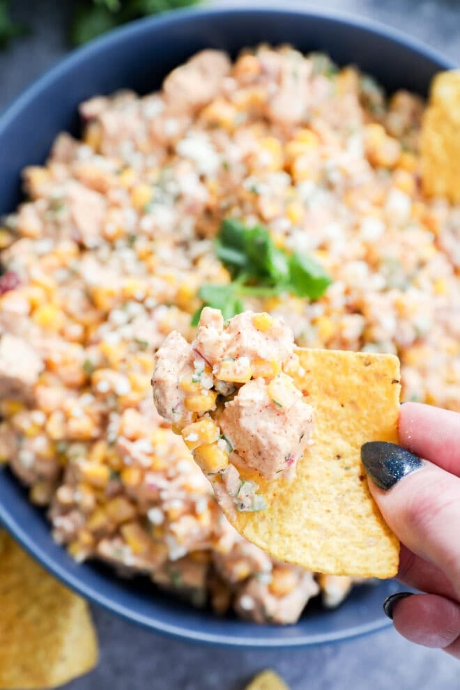 A hand holding a tortilla chip topped with creamy mexican chicken salad—diced chicken, corn, and herbs—above a bowl filled with more of the delicious dip.