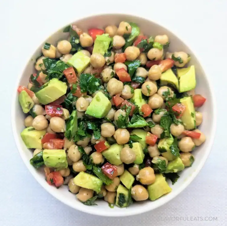 A white bowl filled with a colorful salad made of chickpeas, diced avocado, red bell pepper, chopped parsley, and cilantro on a light background.