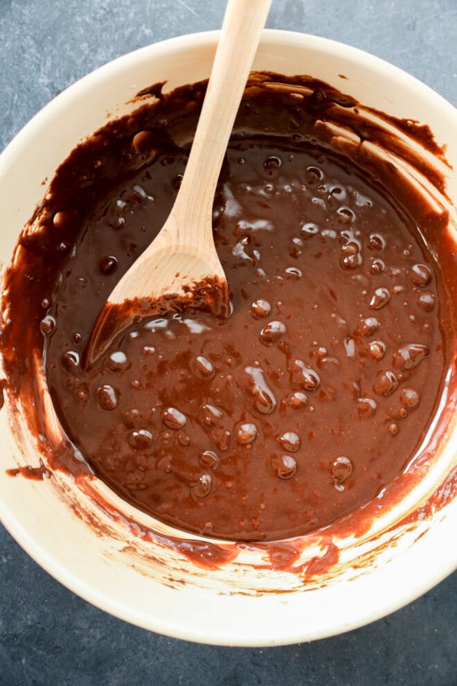 A bowl of brownie batter with a wooden spoon resting inside. The batter is thick and glossy with some air bubbles visible on the surface. The bowl sits on a dark countertop.