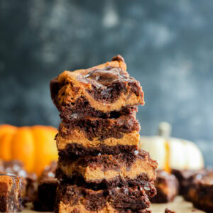 A stack of four swirled chocolate and pumpkin bars sits on parchment paper, with chocolate chips scattered nearby and blurred pumpkins in the background.
