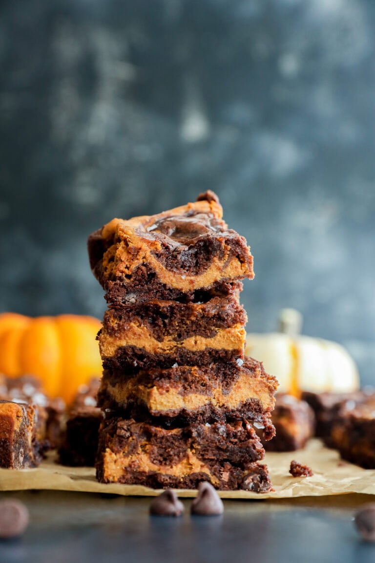 A stack of four swirled chocolate and pumpkin bars sits on parchment paper, with chocolate chips scattered nearby and blurred pumpkins in the background.
