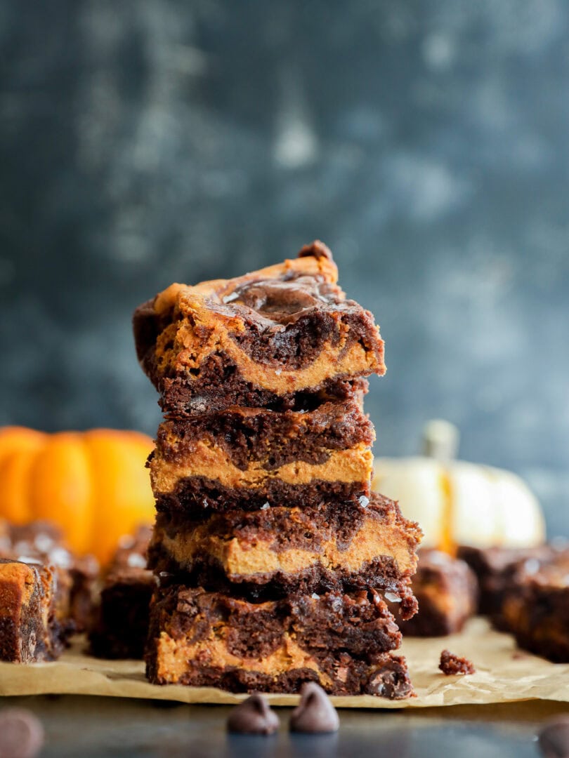 A stack of four swirled chocolate and pumpkin bars sits on parchment paper, with chocolate chips scattered nearby and blurred pumpkins in the background.