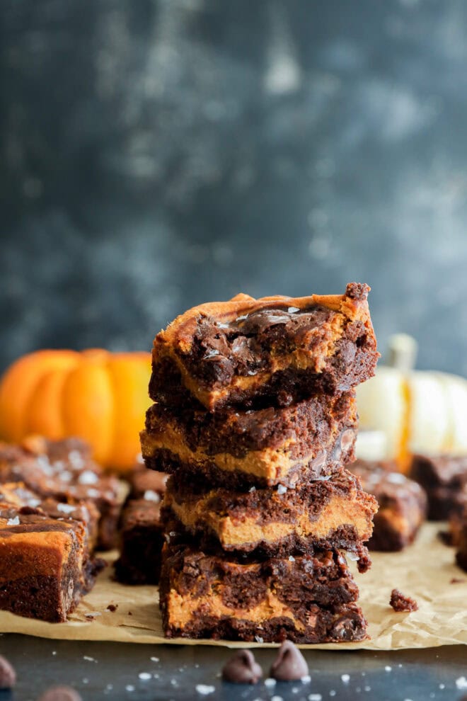 A stack of four pumpkin chocolate chip brownies sits on parchment paper, with a bite taken out of the top brownie. In the background, there are pumpkins and more brownies, set against a dark, blurred backdrop.