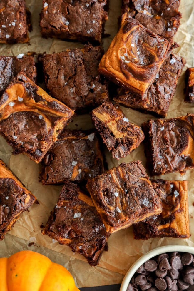 A batch of swirled brownies topped with flaky sea salt sits on parchment paper. Some brownies are stacked, revealing fudgy centers. A bowl of chocolate chips and a small pumpkin are in the corner.