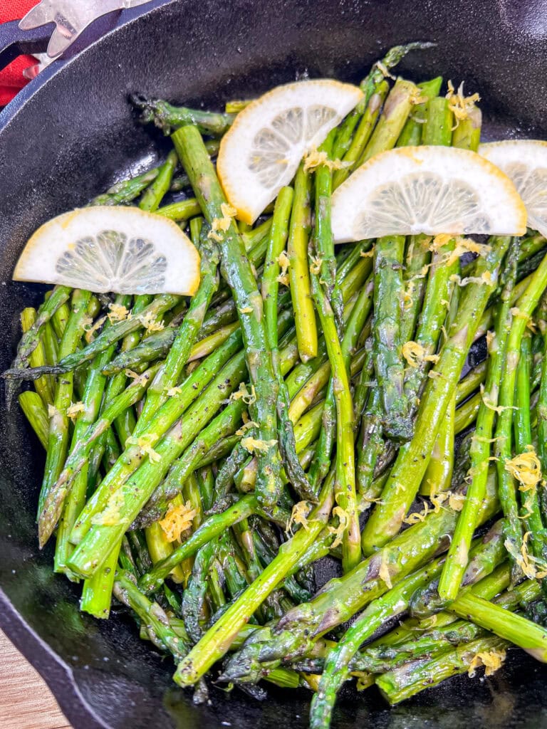 A cast iron skillet filled with cooked asparagus spears, topped with lemon zest and garnished with four thin lemon slices.