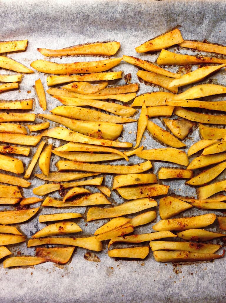 Golden, seasoned potato fries are spread out on a parchment-lined baking tray, evenly spaced and baked to a crisp, with some fries showing light browning.