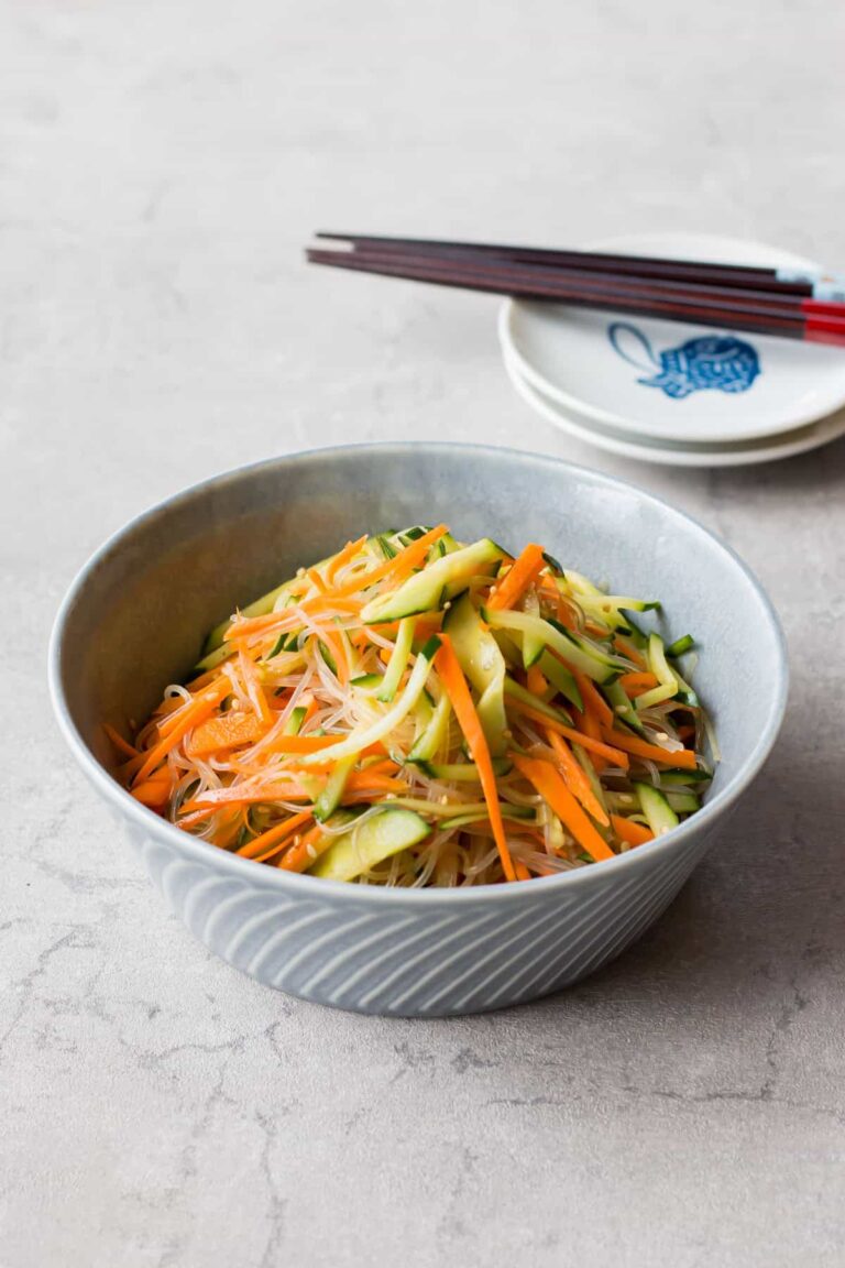 A bowl of glass noodle salad with julienned carrots and cucumbers sits on a light table, with chopsticks resting on a small dish in the background.