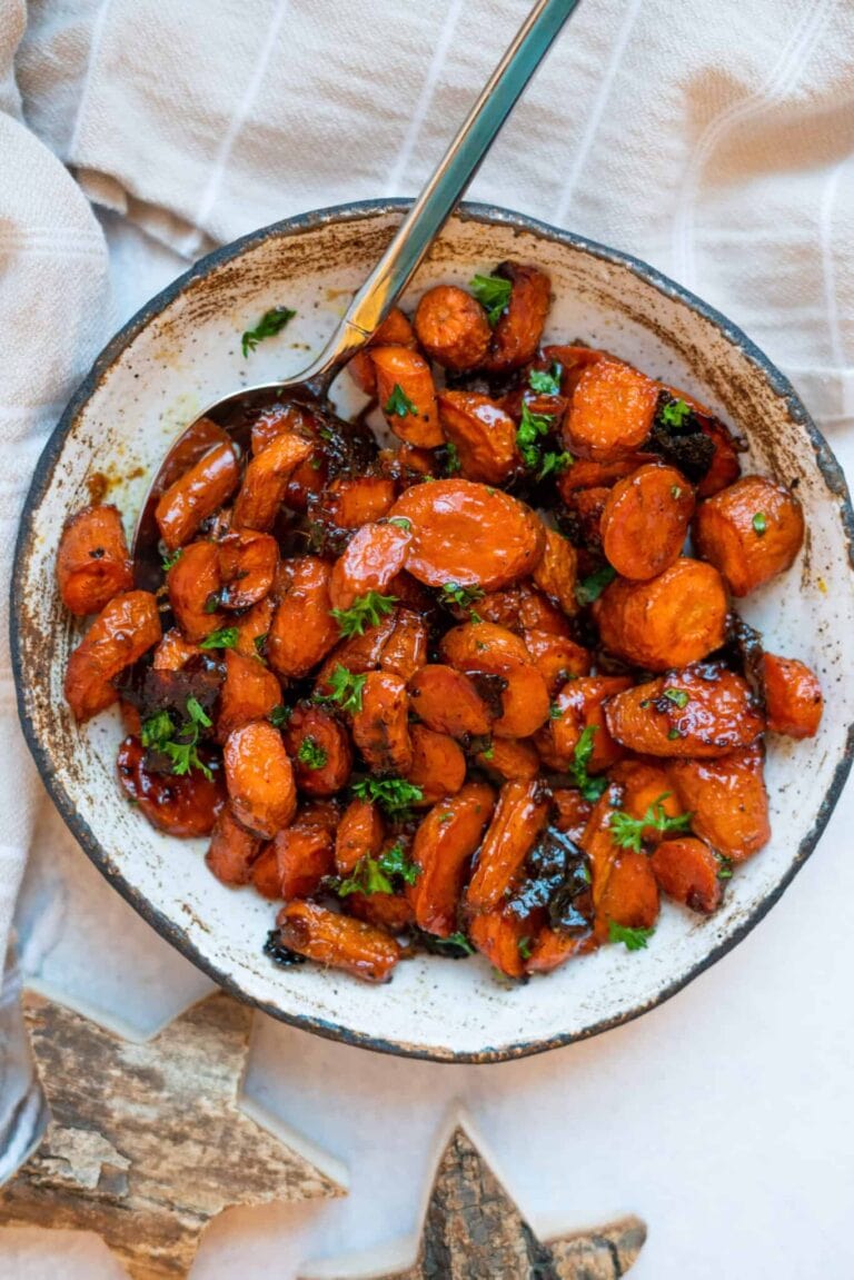 A bowl of glazed roasted carrots garnished with chopped parsley, served with a spoon on a white surface next to a striped cloth napkin.