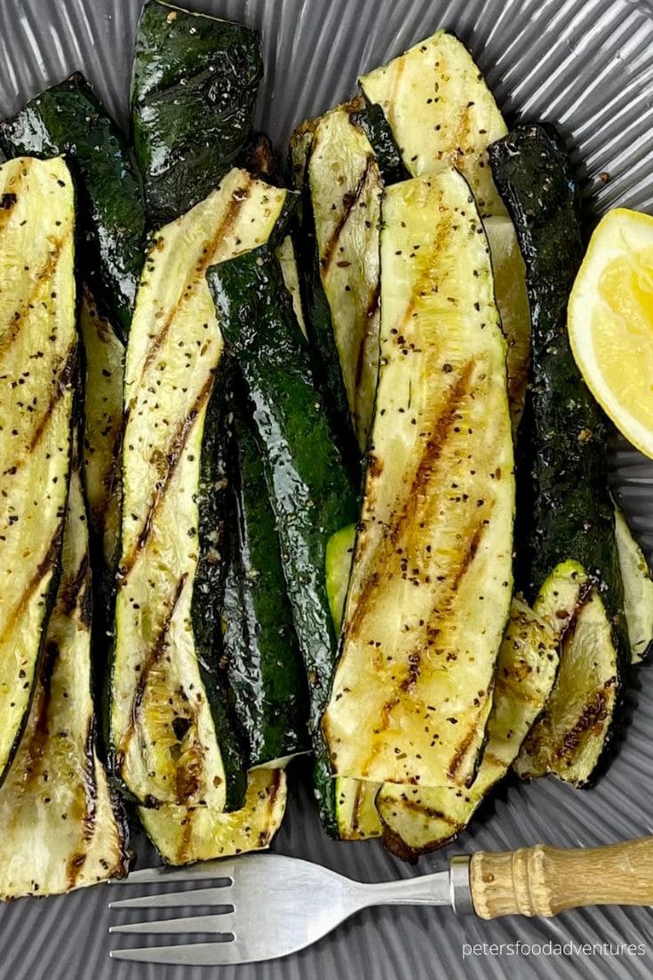 Grilled zucchini slices with char marks, seasoned with pepper, arranged on a dark plate next to a lemon wedge. A silver fork is placed at the bottom of the plate.
