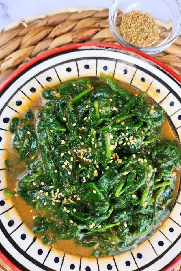 A bowl of seasoned spinach topped with sesame seeds, served on a patterned plate with a small glass dish of sesame seeds in the background.