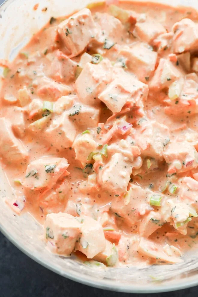 Close-up of a creamy, orange-tinted salad with chunks of chicken, diced vegetables, and chopped herbs mixed together in a bowl.