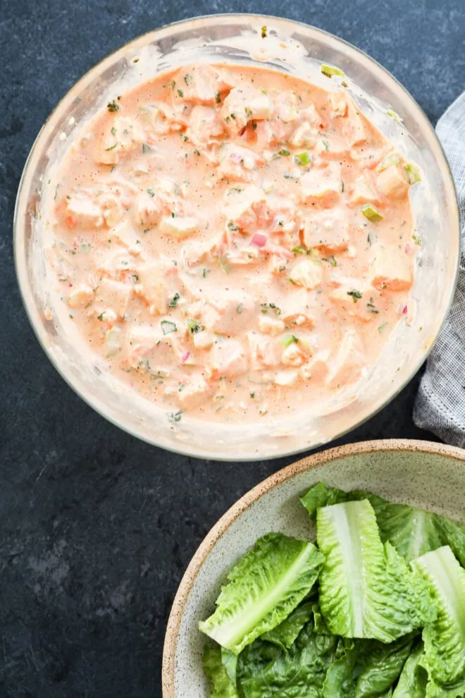 A glass bowl filled with a creamy mixture of chopped vegetables and herbs next to a ceramic bowl containing fresh romaine lettuce leaves, both on a dark tabletop.