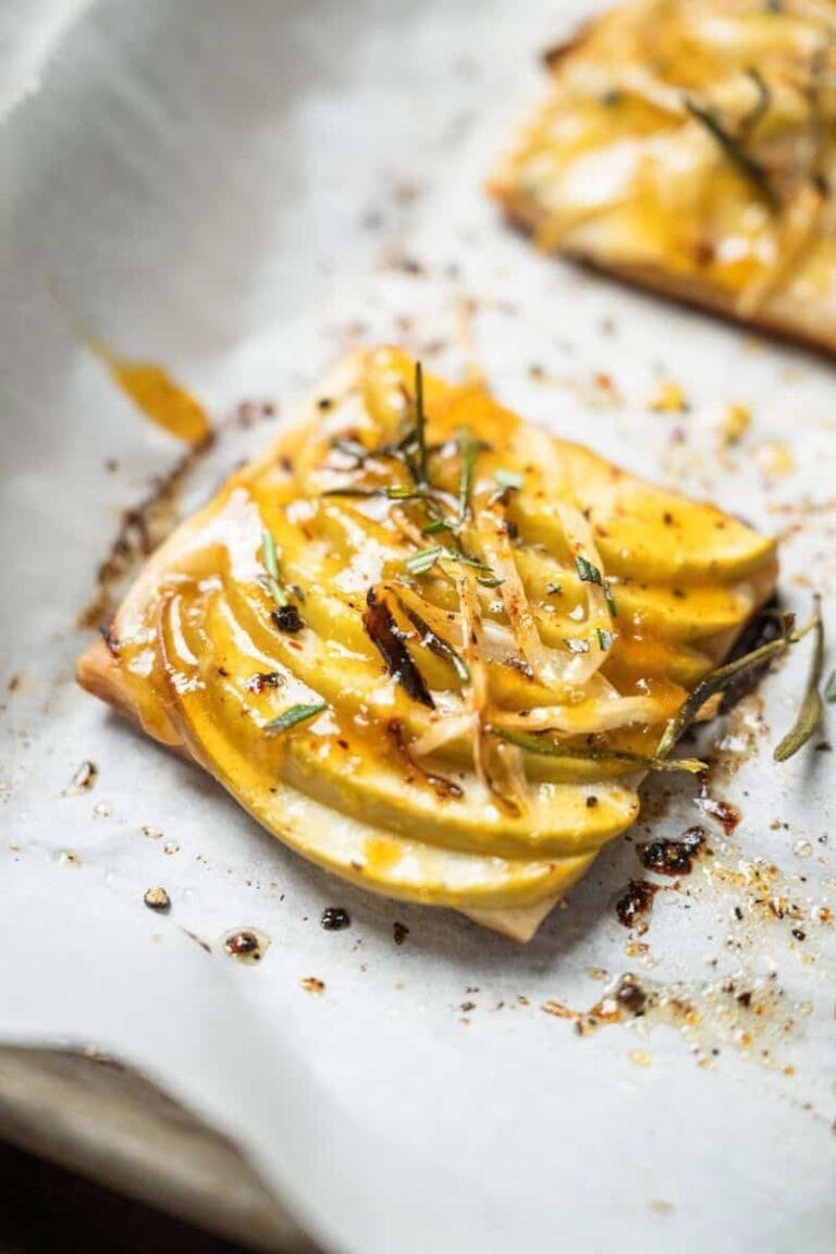 A close-up of a rectangular apple tart made with puff pastry, topped with thinly sliced apples, caramelized onions, herbs, and a golden glaze, sitting on parchment paper—an elegant twist on classic puff pastry appetizers.