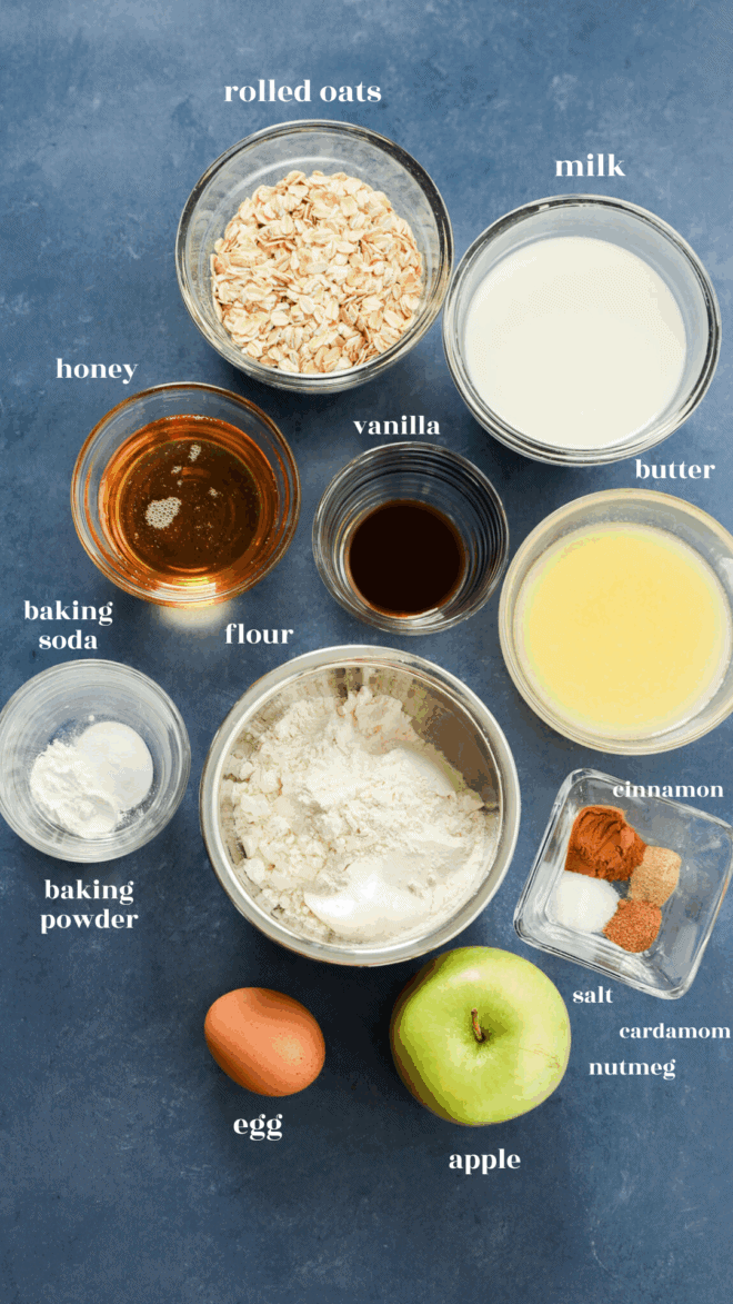 A flat lay of baking ingredients for apple oatmeal muffins on a blue surface, including rolled oats, milk, butter, vanilla, honey, flour, baking soda, cinnamon, spices, salt, an egg, and a green apple.
