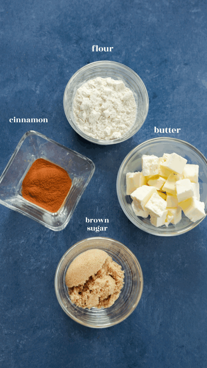 Four small bowls on a blue surface, each labeled: flour (white powder), butter (cubed), brown sugar (granulated light brown), and cinnamon—classic ingredients for delicious apple oatmeal muffins.