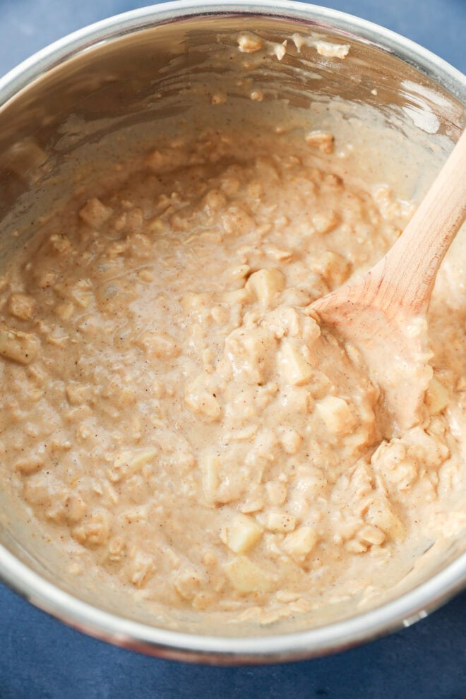A metal mixing bowl filled with thick, creamy batter for apple oatmeal muffins, packed with chopped apples and oats, being stirred with a wooden spoon.