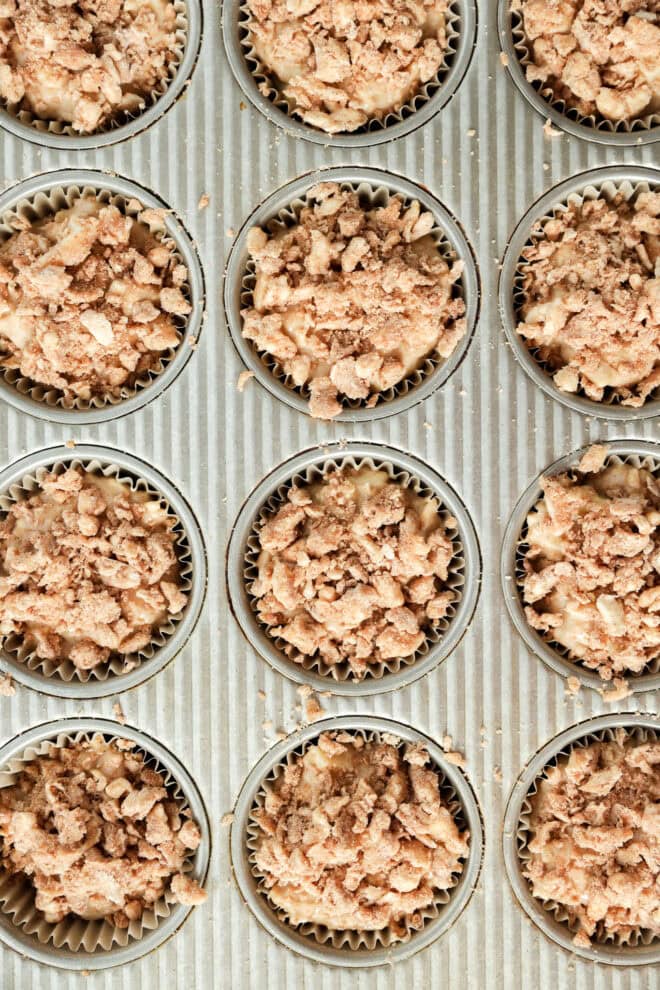 A muffin tin filled with paper liners, each containing unbaked apple oatmeal muffins batter topped with a crumbly streusel topping, viewed from above.