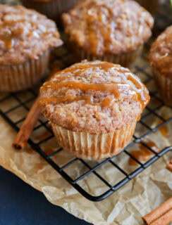 A close-up of cinnamon and apple oatmeal muffins drizzled with caramel sauce on a wire rack, with parchment paper and cinnamon sticks nearby.