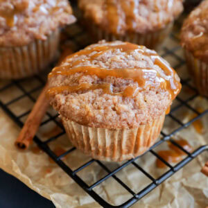 A close-up of cinnamon and apple oatmeal muffins drizzled with caramel sauce on a wire rack, with parchment paper and cinnamon sticks nearby.