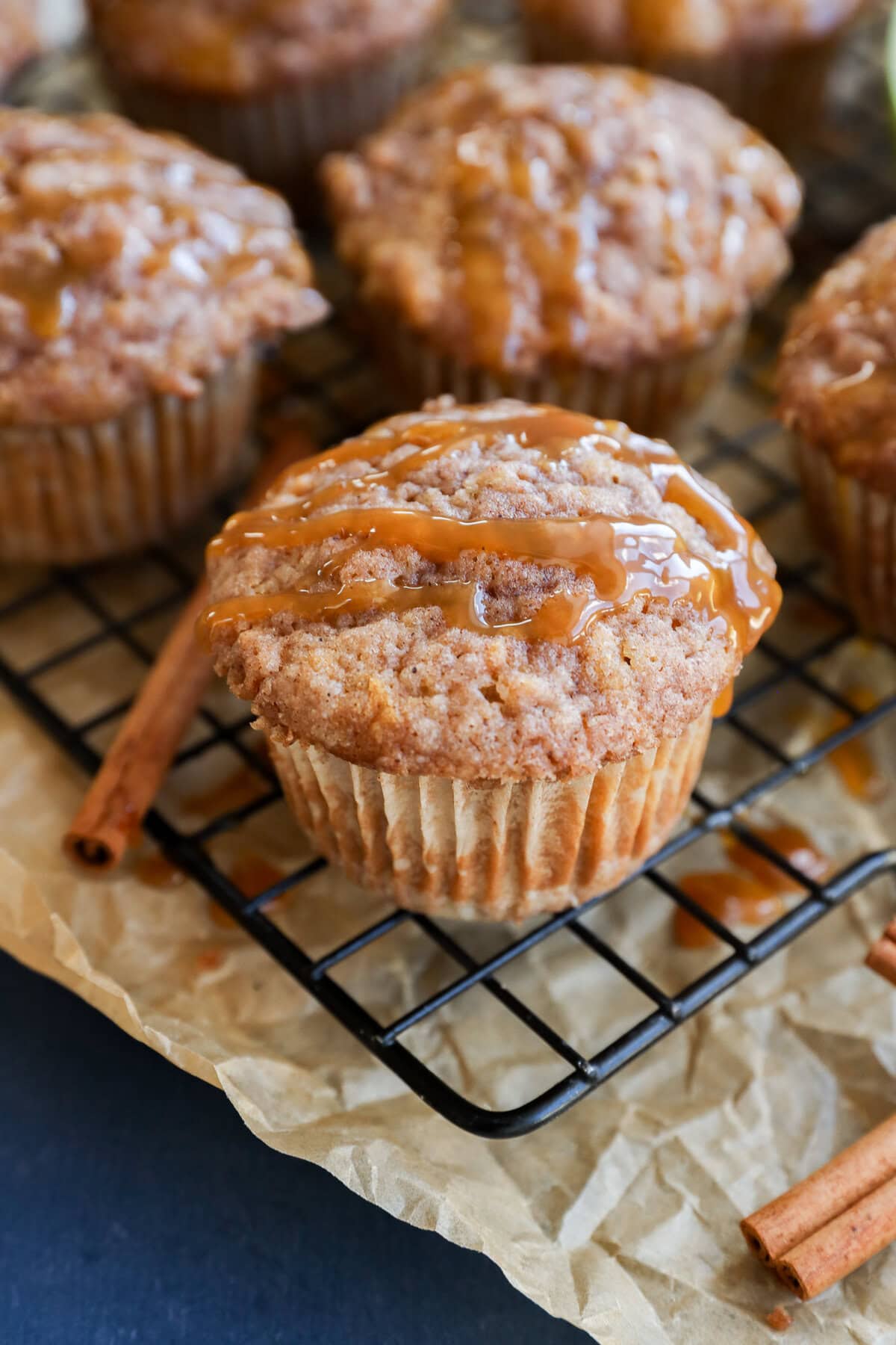 A close-up of cinnamon and apple oatmeal muffins drizzled with caramel sauce on a wire rack, with parchment paper and cinnamon sticks nearby.
