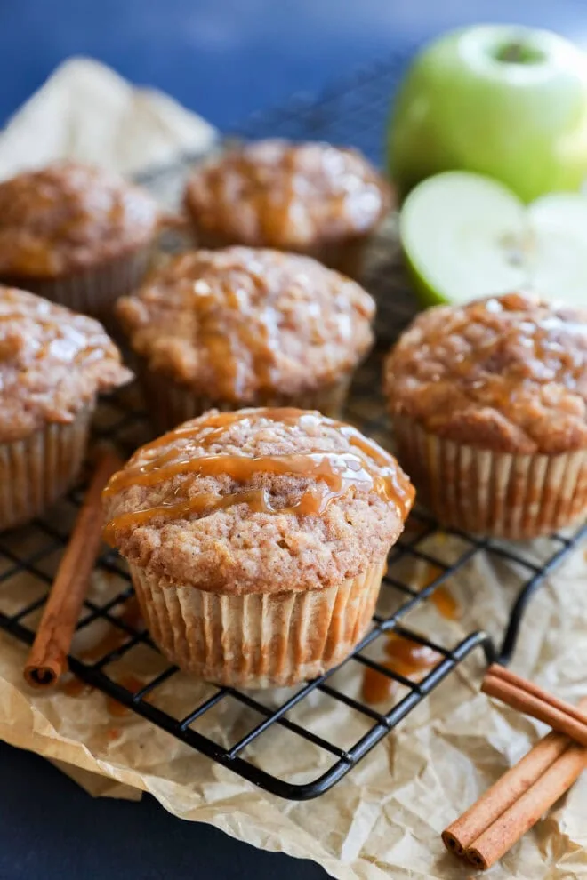 Freshly baked apple oatmeal muffins, drizzled with caramel, sit on a cooling rack lined with parchment paper. Cinnamon sticks and a green apple, both whole and halved, are visible in the background.