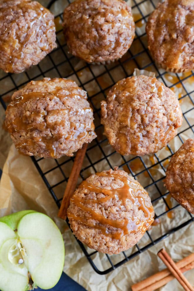 Apple oatmeal muffins topped with crumbly streusel and caramel drizzle are cooling on a wire rack. Cinnamon sticks and a sliced green apple rest nearby on parchment paper, completing this cozy scene.