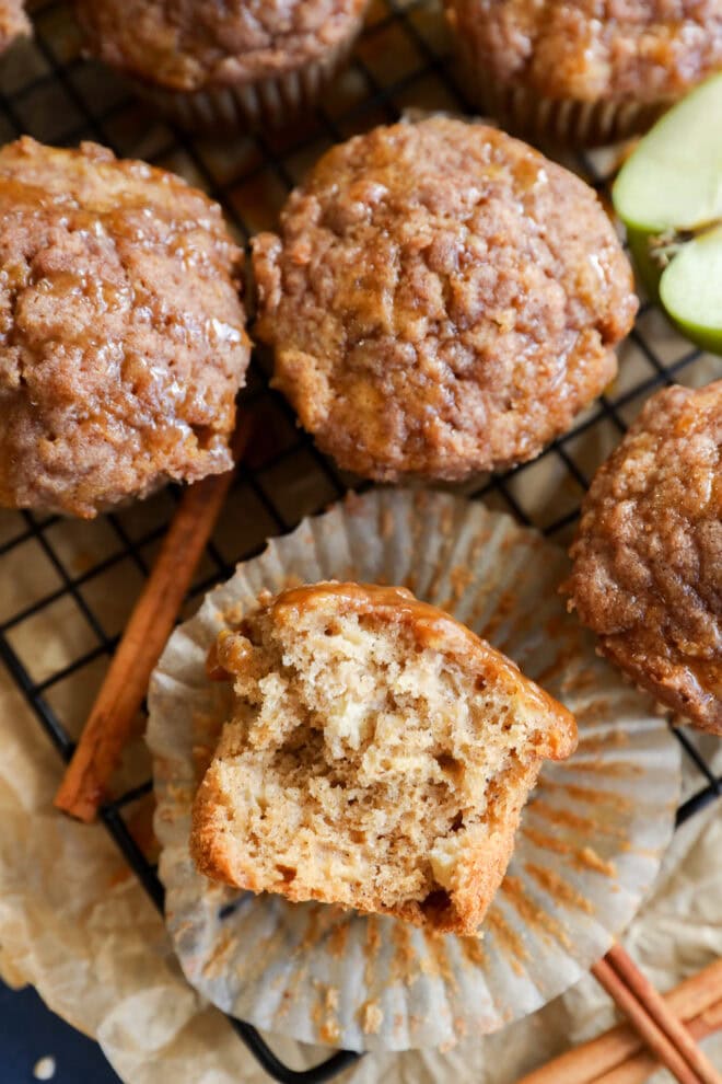 A close-up of apple oatmeal muffins on a wire rack, one muffin partially unwrapped with a bite taken, revealing a moist, fluffy interior. Cinnamon sticks and apple slices are nearby.