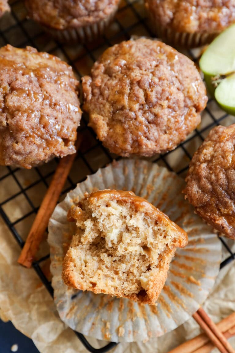 A close-up of apple oatmeal muffins on a wire rack, one muffin partially unwrapped with a bite taken, revealing a moist, fluffy interior. Cinnamon sticks and apple slices are nearby.