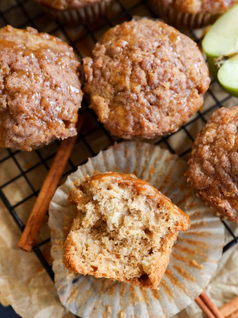 A close-up of apple oatmeal muffins on a wire rack, one muffin partially unwrapped with a bite taken, revealing a moist, fluffy interior. Cinnamon sticks and apple slices are nearby.