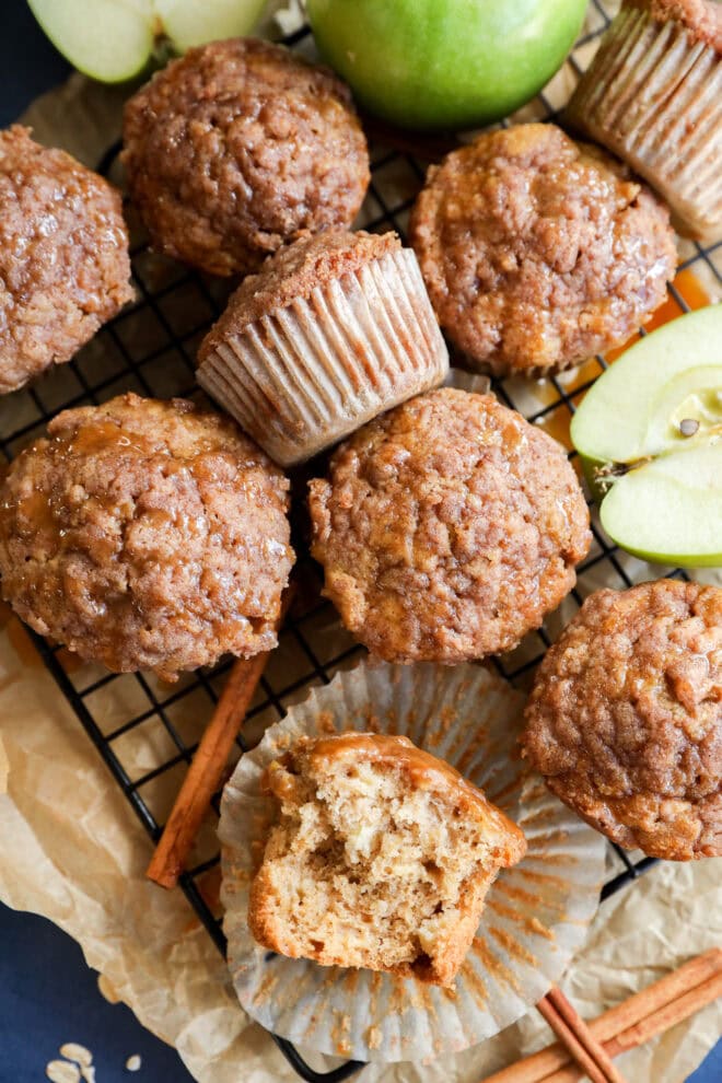 A wire rack holds several apple oatmeal muffins, some with paper liners. One muffin is cut open, showing a moist interior. Sliced green apples and cinnamon sticks rest on crumpled parchment paper nearby.