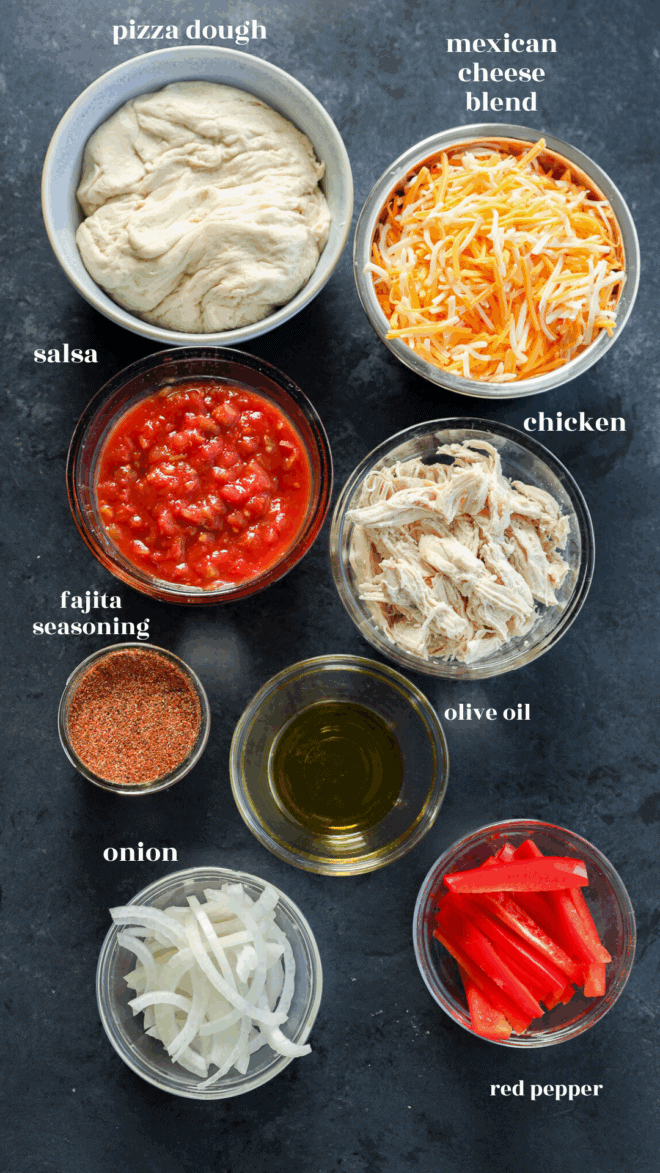 Overhead view of ingredients in bowls: pizza dough, shredded Mexican cheese blend, salsa, shredded chicken, fajita seasoning, olive oil, sliced onion, and sliced red pepper on a dark surface.