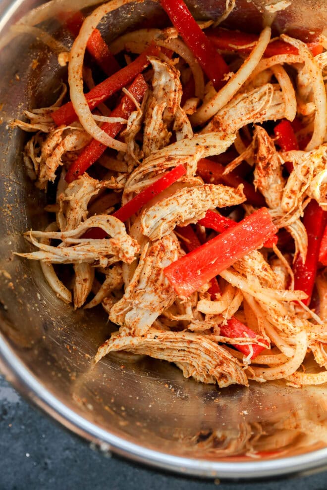 Shredded chicken mixed with sliced red bell peppers and onions, all seasoned with spices, in a metal bowl.