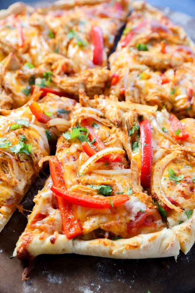Close-up of a sliced pizza topped with melted cheese, shredded chicken, red bell pepper strips, onions, and fresh cilantro on a thick, crispy crust.
