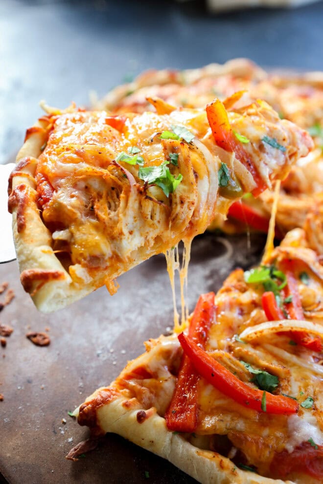 A close-up of a chicken fajita pizza slice being lifted, topped with melted cheese, chicken, red bell peppers, onions, and sprinkled with fresh herbs. The crust looks golden and crispy, with cheese stretching as the slice is pulled away.