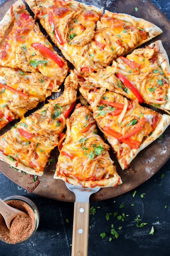 A fresh, homemade pizza topped with shredded chicken, red bell peppers, melted cheese, and herbs sits on a round pizza stone. One slice is being lifted with a wooden-handled spatula. A small bowl of seasoning is nearby.