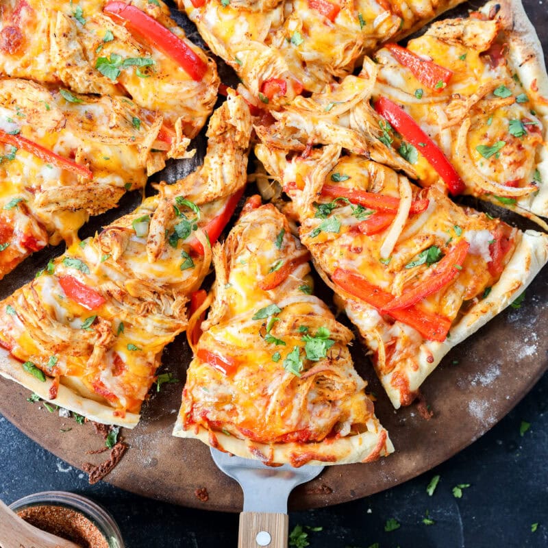 A fresh, homemade pizza topped with shredded chicken, red bell peppers, melted cheese, and herbs sits on a round pizza stone. One slice is being lifted with a wooden-handled spatula. A small bowl of seasoning is nearby.