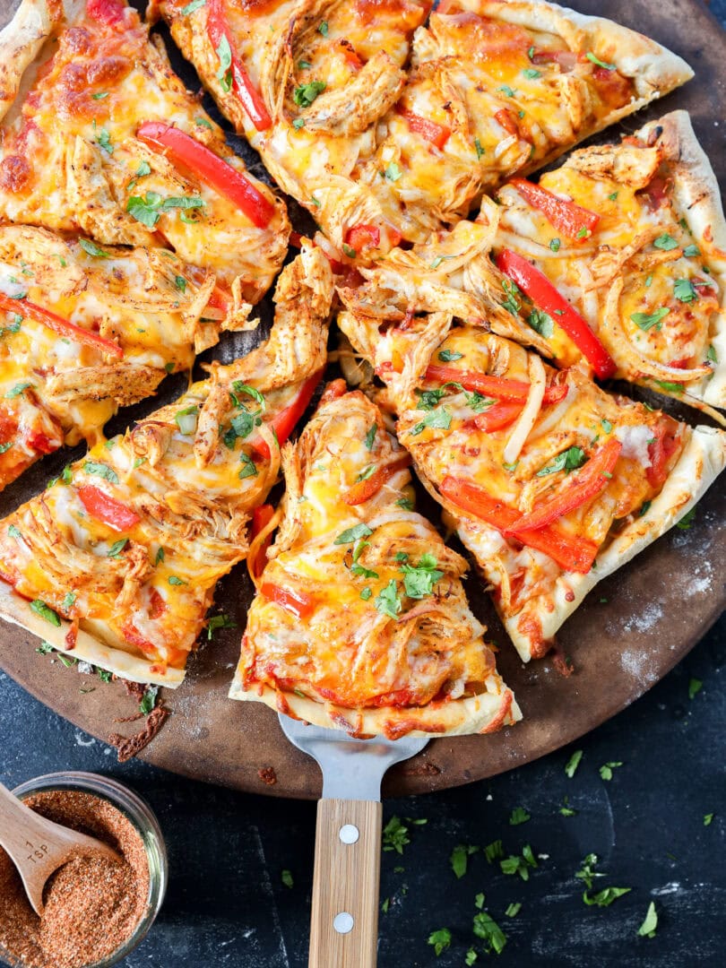 A fresh, homemade pizza topped with shredded chicken, red bell peppers, melted cheese, and herbs sits on a round pizza stone. One slice is being lifted with a wooden-handled spatula. A small bowl of seasoning is nearby.