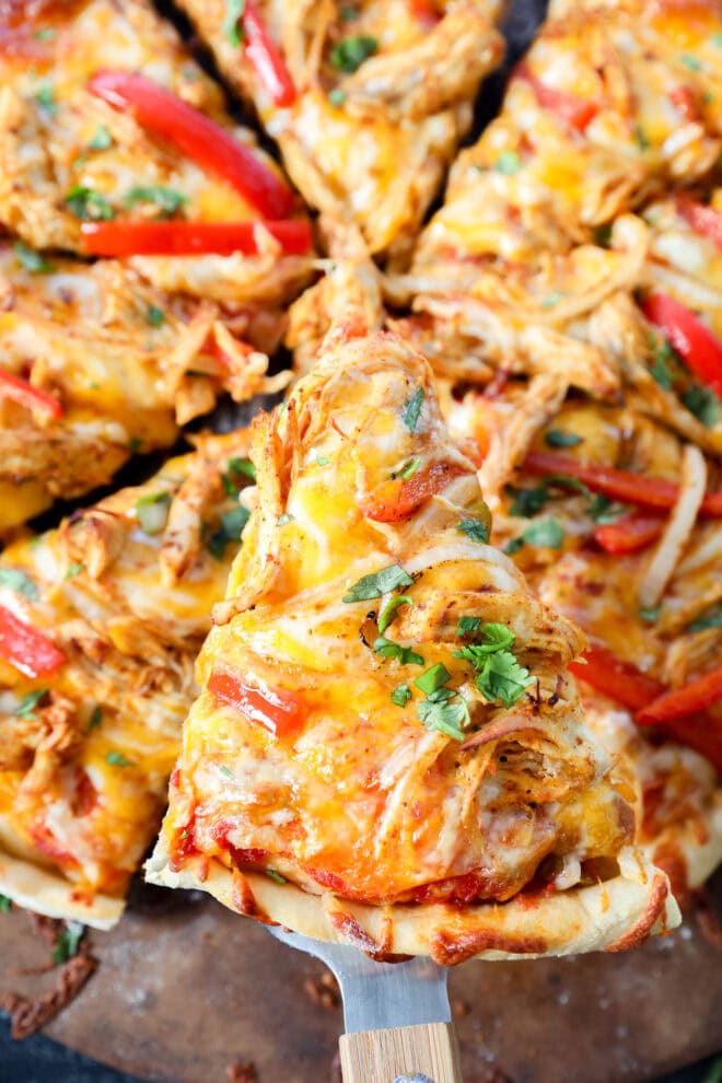 A close-up of a pizza topped with shredded chicken, melted cheese, red bell peppers, onions, and fresh herbs. One slice is being lifted from the whole chicken fajita pizza with a spatula.