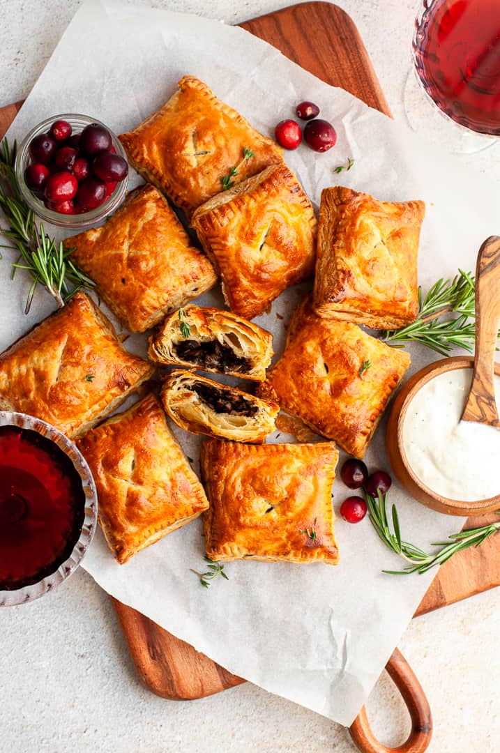 Golden, flaky puff pastry appetizers are arranged on parchment paper with a bowl of cranberries, a glass of red drink, a bowl of white dipping sauce, and fresh rosemary sprigs, all atop a wooden board.